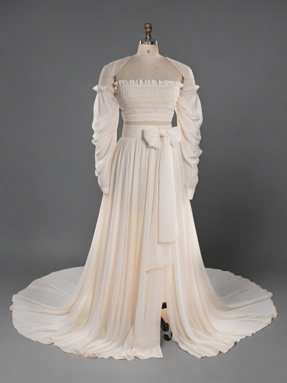An off-the-shoulder cream-colored gown displayed on a dress form mannequin against a solid gray studio background. The dress features a smocked bodice, long voluminous sleeves, a matching waist sash, and a long train.