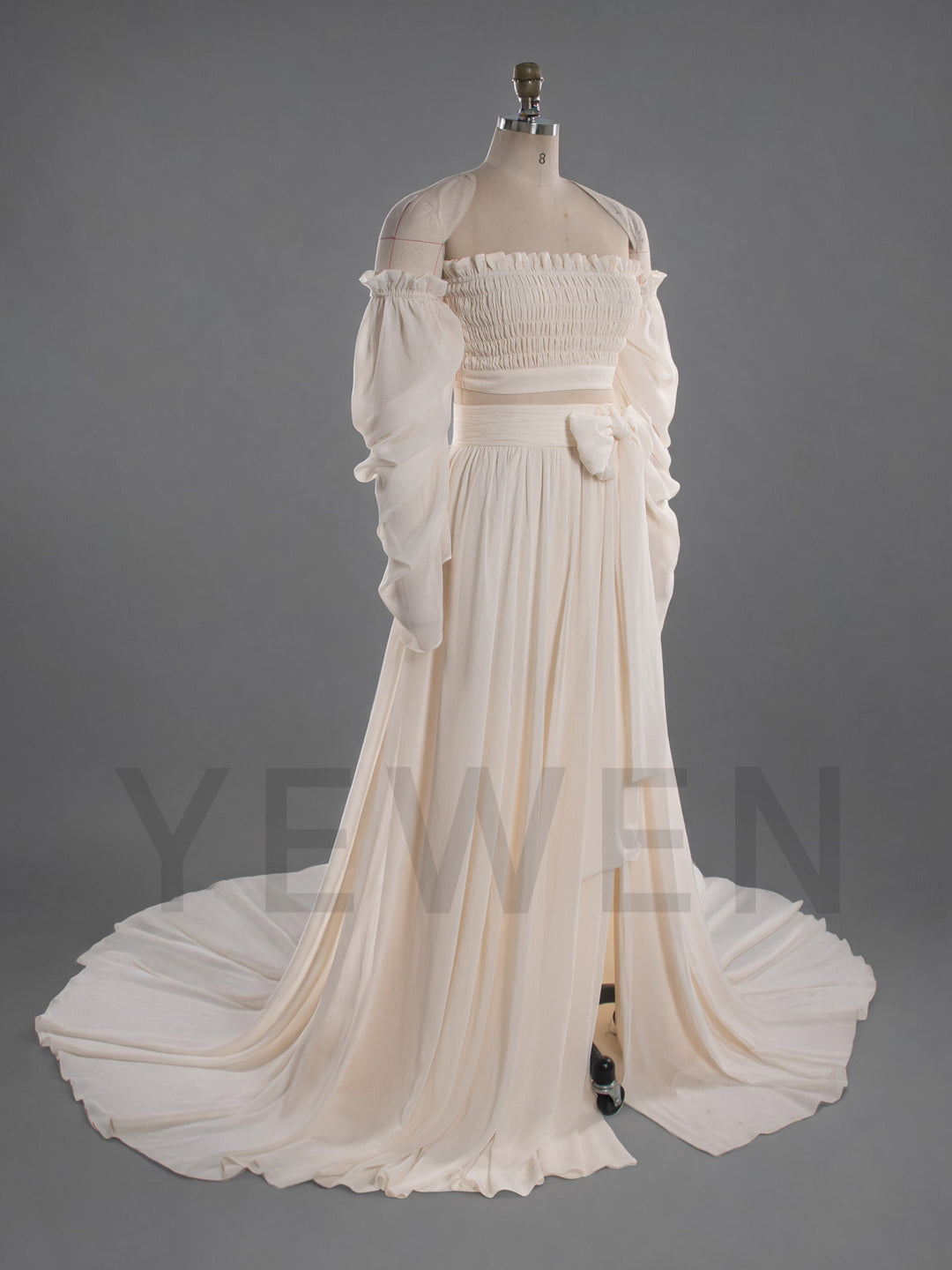 An off-the-shoulder, floor-length cream dress displayed on a mannequin against a solid gray studio background. The dress features a smocked bodice, long voluminous sleeves, a matching waist sash with a bow, and a full skirt with a slight train.