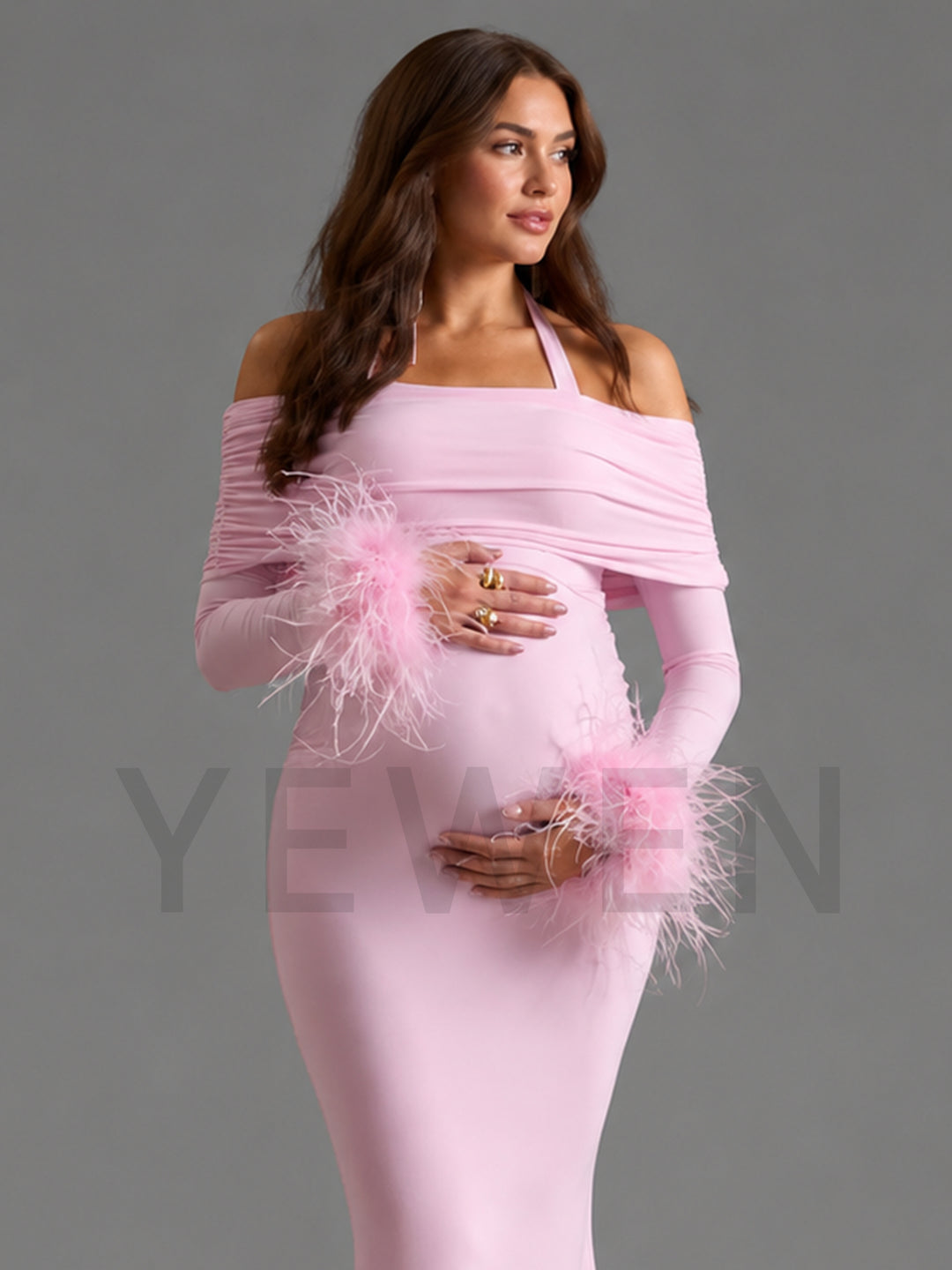 A pregnant model wearing a light pink off-the-shoulder maternity dress with long sleeves and fluffy feather trim at the cuffs, posed against a solid gray studio background.