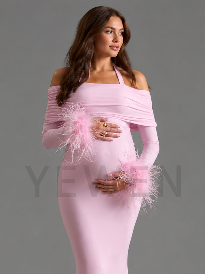 A pregnant model wearing a light pink off-the-shoulder maternity dress with long sleeves and fluffy feather trim at the cuffs, posed against a solid gray studio background.