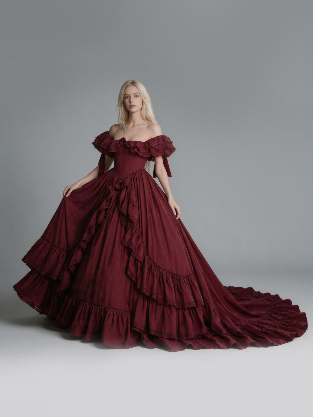 A model wearing a voluminous off-the-shoulder ball gown in deep burgundy, set against a solid gray studio background.