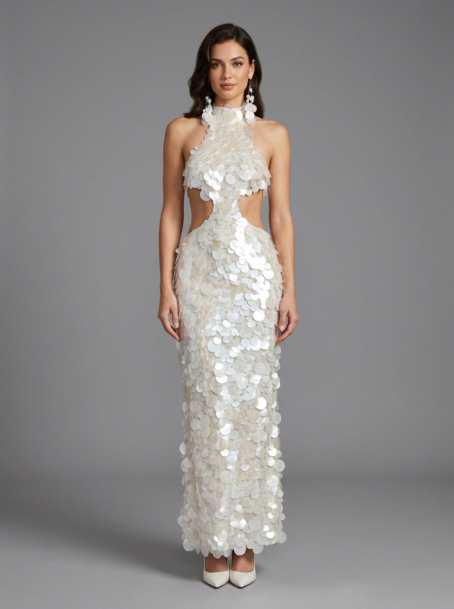 A woman modeling a floor-length white halter gown covered in layered shell-like embellishments, with side cutouts and a fitted silhouette, against a plain gray studio background.