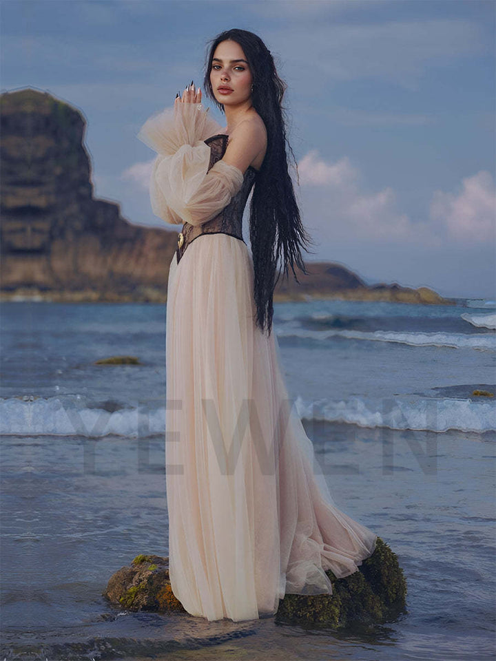 Woman in a long, flowing dress standing on a rocky beach with ocean waves and cliffs in the background.