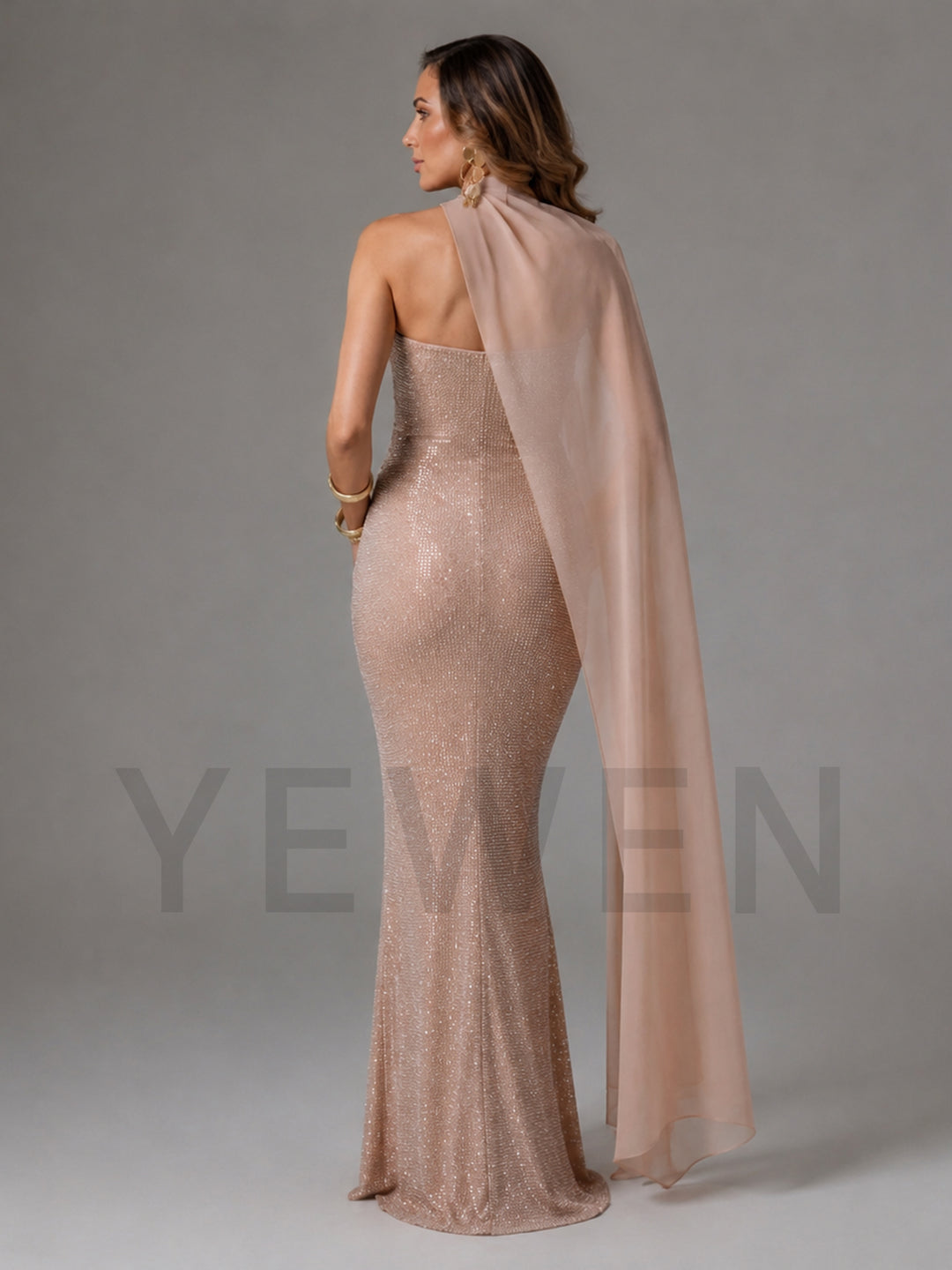 A woman modeling a floor-length, strapless champagne beige sequined gown with a matching sheer draped cape, shown from the back against a neutral gray studio background.