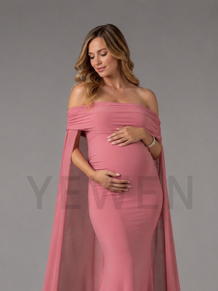 A pregnant woman wearing a dusty pink off-the-shoulder maternity gown with a flowing sheer cape, posing against a solid gray studio background.