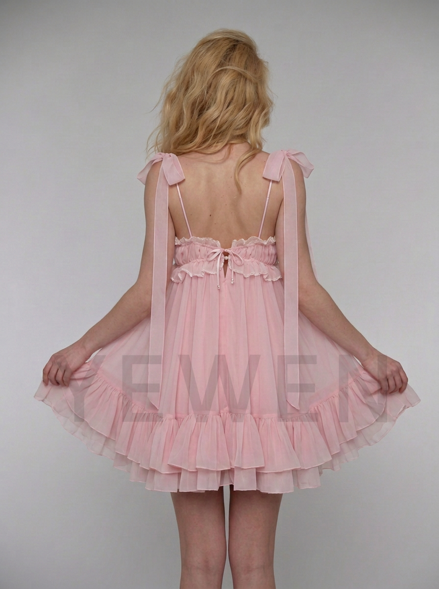 A woman shown from behind wearing a light pink ruffled nightgown with tie-shoulder straps and a ruffled tulle skirt, standing against a plain light gray studio background.