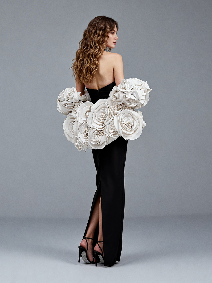 Back view of a model wearing a black elegant evening dress with a detachable 3D white floral cape and back slit.