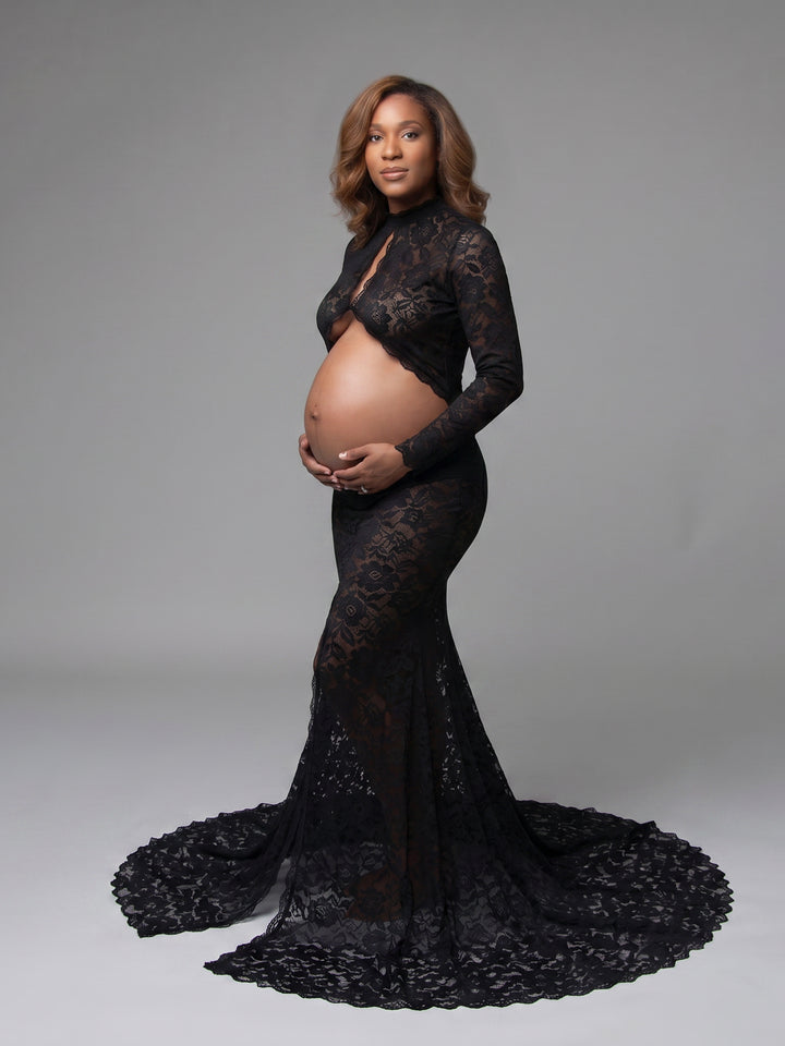 A pregnant model wearing a black lace maternity gown with long sleeves, a high neckline, and a dramatic train, standing against a solid gray studio background.
