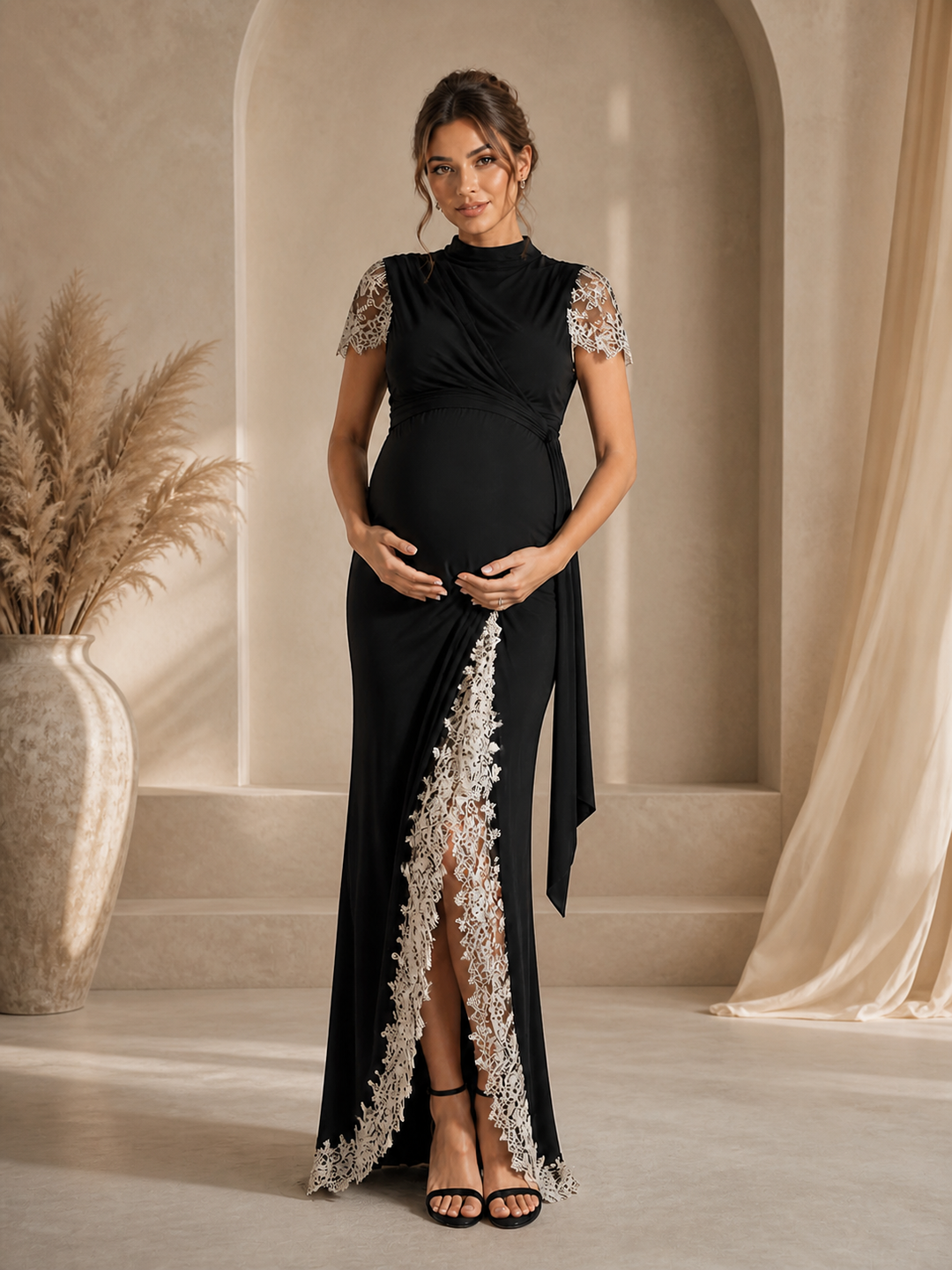 A pregnant woman models a black maternity maxi gown with silver lace detailing on the sleeves and a high slit, standing in a warm, neutral-toned room with arches and a pampas grass vase.