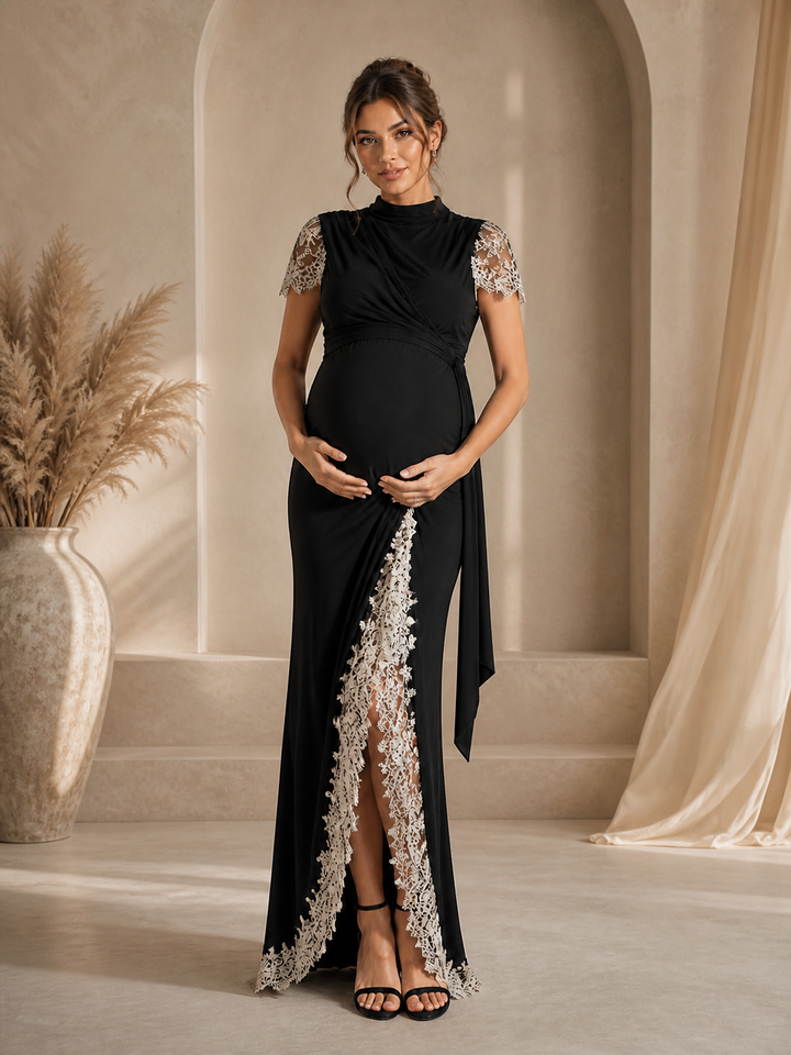 A pregnant woman models a black maternity maxi gown with silver lace detailing on the sleeves and a high slit, standing in a warm, neutral-toned room with arches and a pampas grass vase.