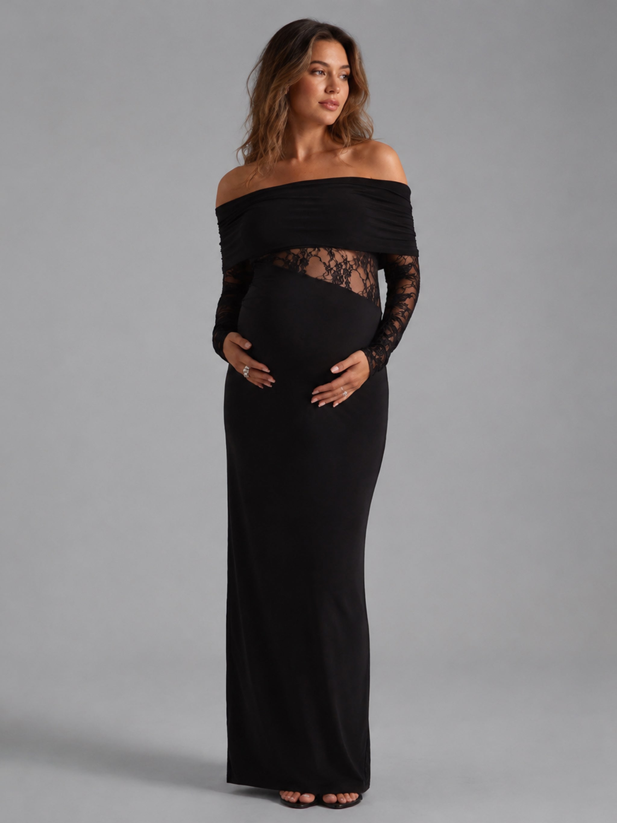 A pregnant woman modeling a black off-the-shoulder maternity maxi dress with long sheer lace sleeves and a lace panel at the waist, photographed against a solid gray studio background.