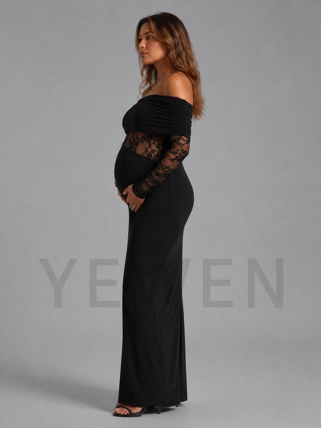 A pregnant woman modeling a black off-the-shoulder maternity maxi dress with long lace sleeves, standing in profile against a solid gray studio background.