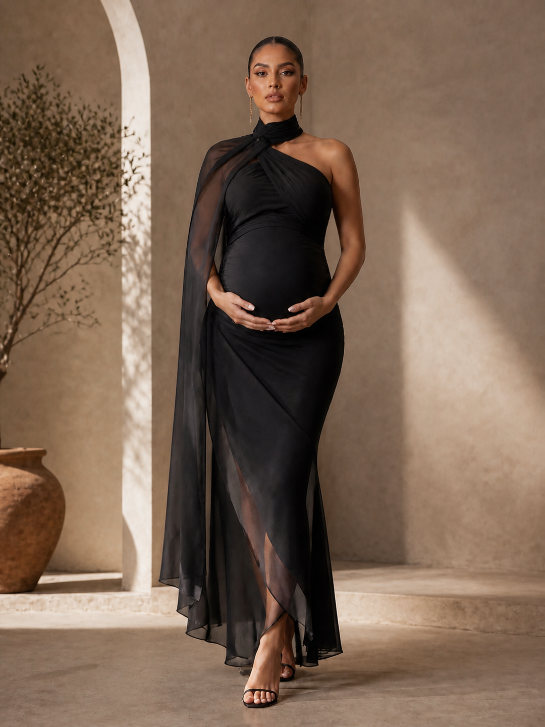 A pregnant model wearing a black one-shoulder draped maternity gown with a sheer overlay and high-low hem, standing in a warm, sunlit architectural setting with beige walls and a large terracotta pot.