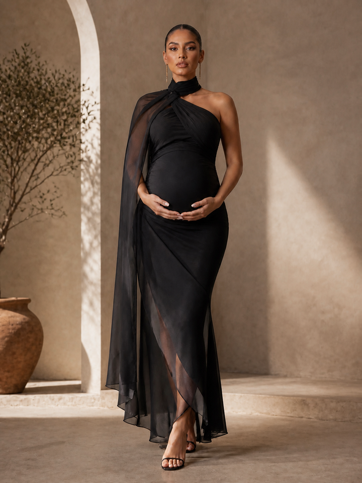 A pregnant model wearing a black one-shoulder draped maternity gown with a sheer overlay and high-low hem, standing in a warm, sunlit architectural setting with beige walls and a large terracotta pot.