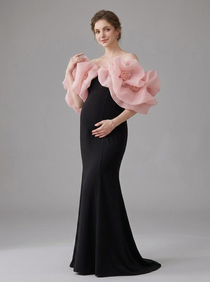 A pregnant model wearing a floor-length black mermaid gown with dramatic oversized light pink fabric flower ruffles on the shoulders, photographed against a neutral gray studio background.