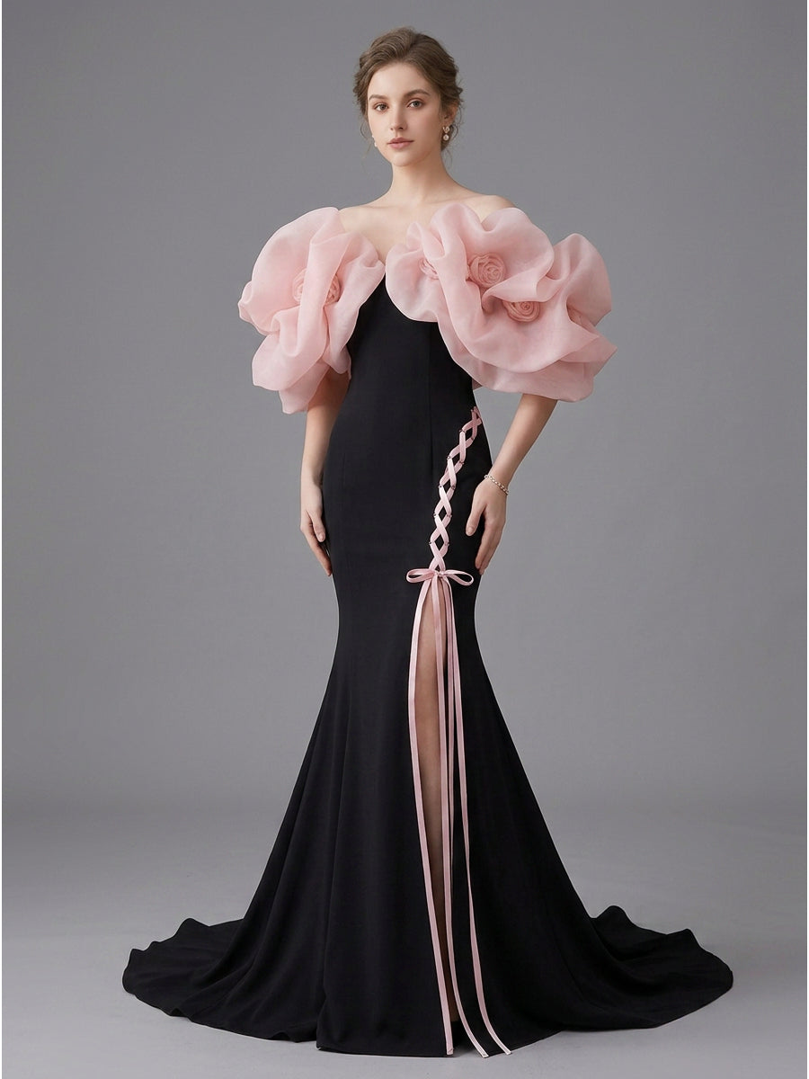 A model wears a floor-length black mermaid gown with dramatic oversized light pink ruffled shoulder details, a pink braided side accent, and a long pink tie detail, set against a solid gray studio background.