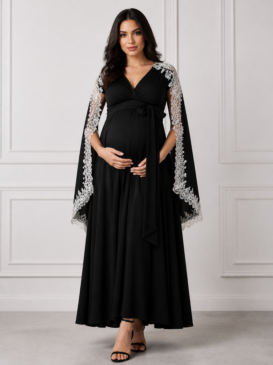 A pregnant woman modeling a black maternity maxi dress with long sheer sleeves featuring white lace trim, standing against a white paneled wall.