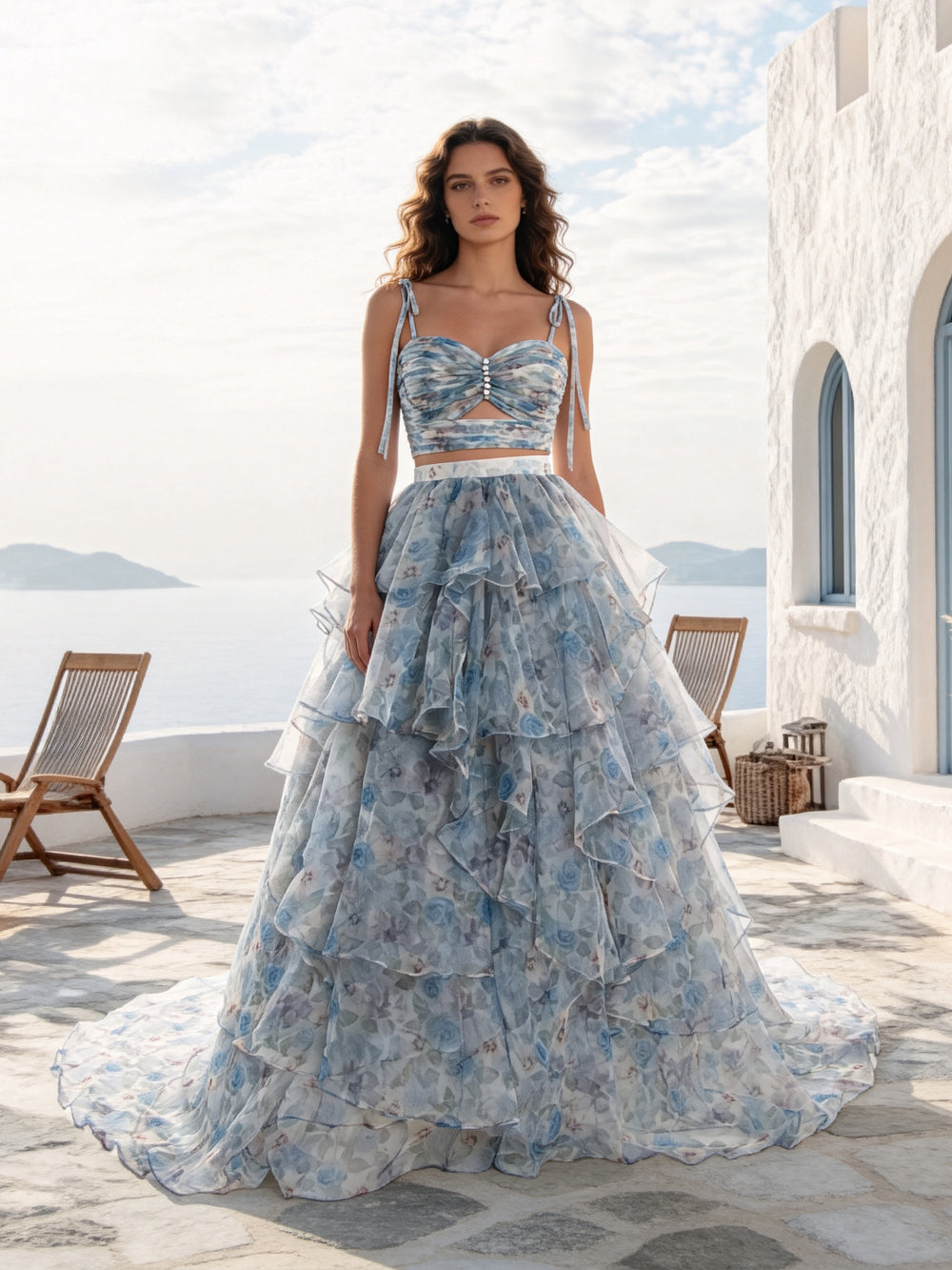 A woman stands on a sunlit stone terrace wearing a voluminous light blue floral gown with a fitted sweetheart-style bodice, thin straps, a waist cutout, and a heavily ruffled tiered skirt. White walls, chairs, and distant water are visible in the background.