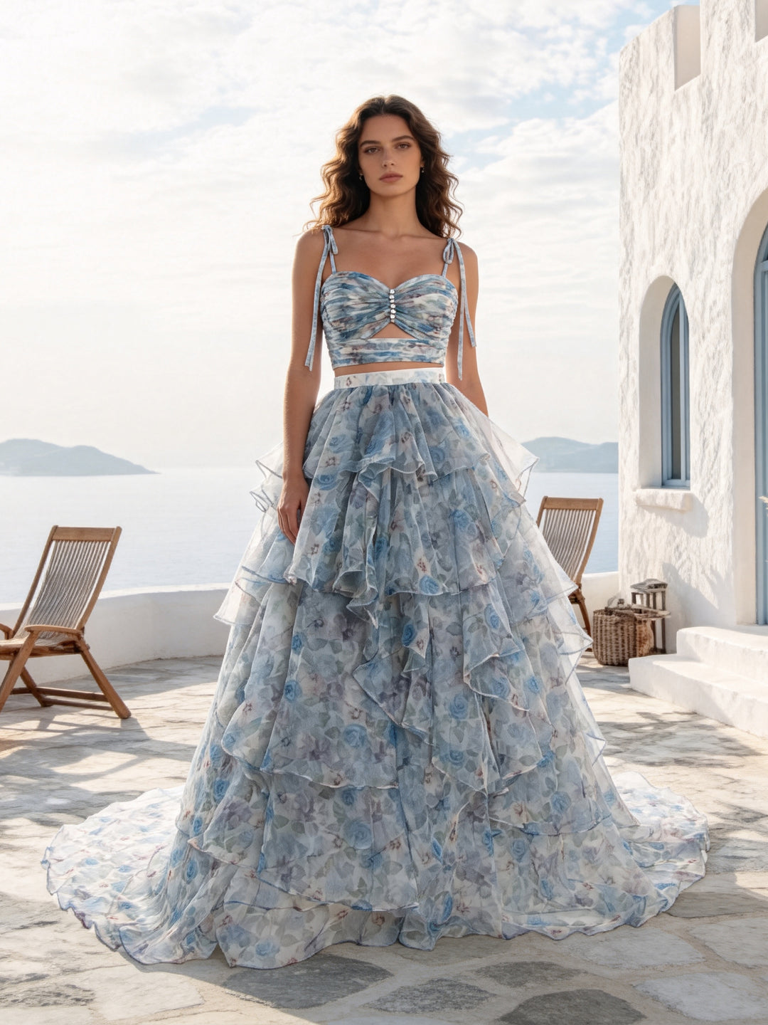 A woman stands on a sunlit stone terrace wearing a voluminous light blue floral gown with a fitted sweetheart-style bodice, thin straps, a waist cutout, and a heavily ruffled tiered skirt. White walls, chairs, and distant water are visible in the background.