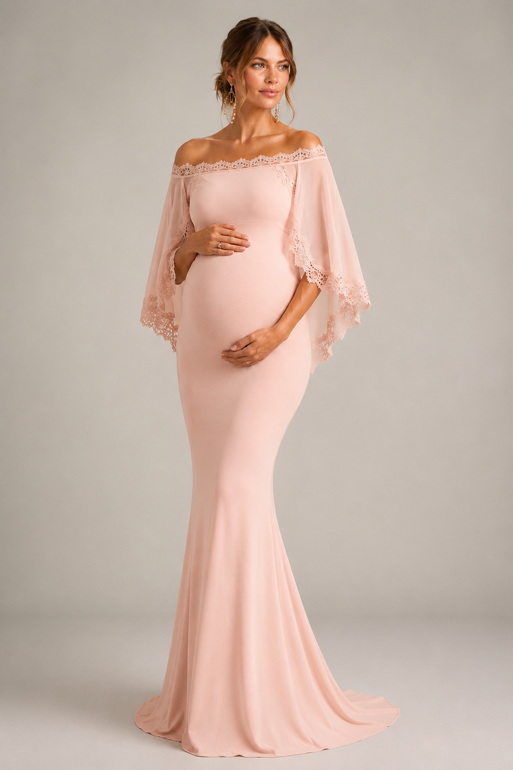 A pregnant model wearing a light blush pink off-the-shoulder maternity gown with sheer flutter sleeves and lace trim, standing against a neutral gray studio background.