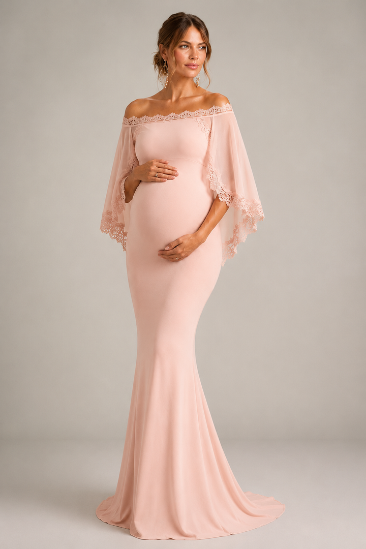 A pregnant model wearing a light blush pink off-the-shoulder maternity gown with sheer flutter sleeves and lace trim, standing against a neutral gray studio background.