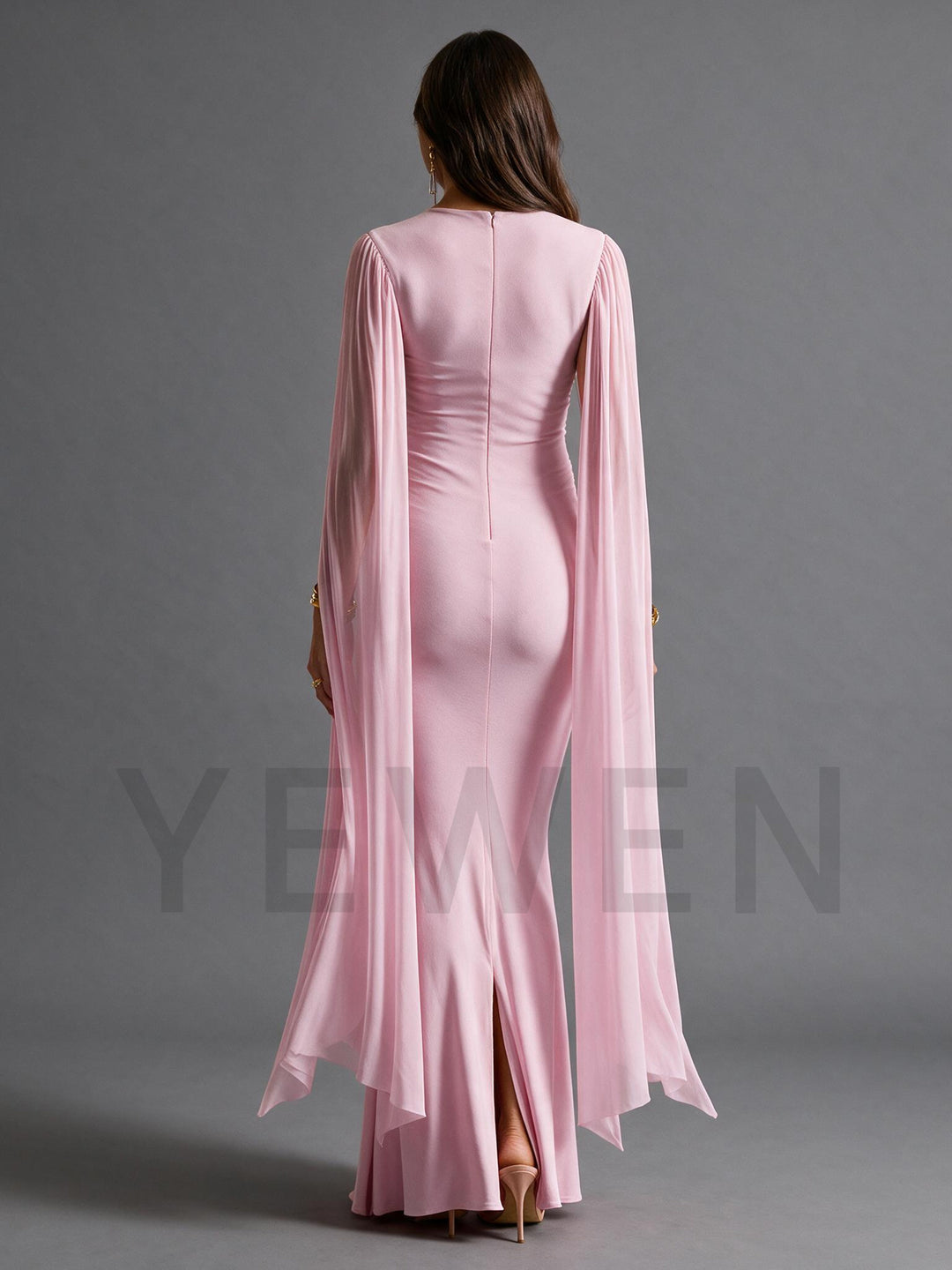 A woman viewed from the back wearing a floor-length light pink gown with long flowing cape sleeves and a fitted silhouette, photographed against a plain gray studio background.