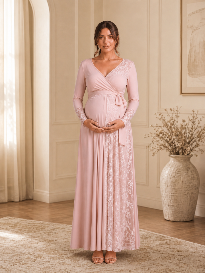 A pregnant woman wearing a long-sleeve, wrap-style maxi maternity dress in light blush pink with floral lace panels, standing in a warm, elegant room with beige walls, a rug, and a large vase.