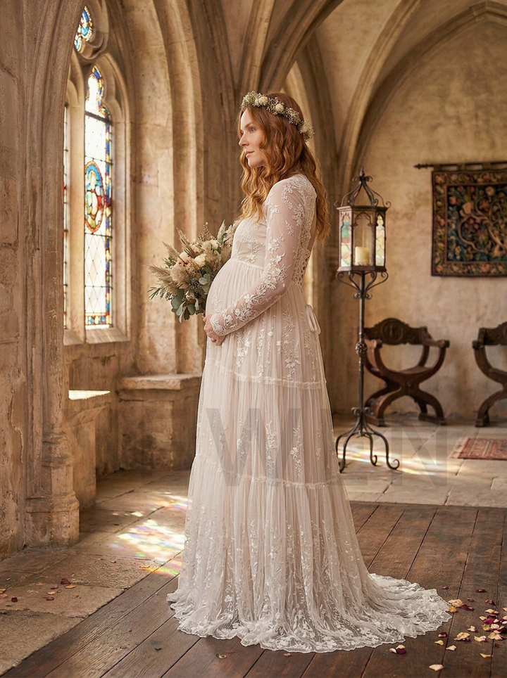 A pregnant woman wearing a long-sleeved, tiered lace wedding gown and a floral crown stands in a stone church hall with stained glass windows, holding a bouquet of flowers.