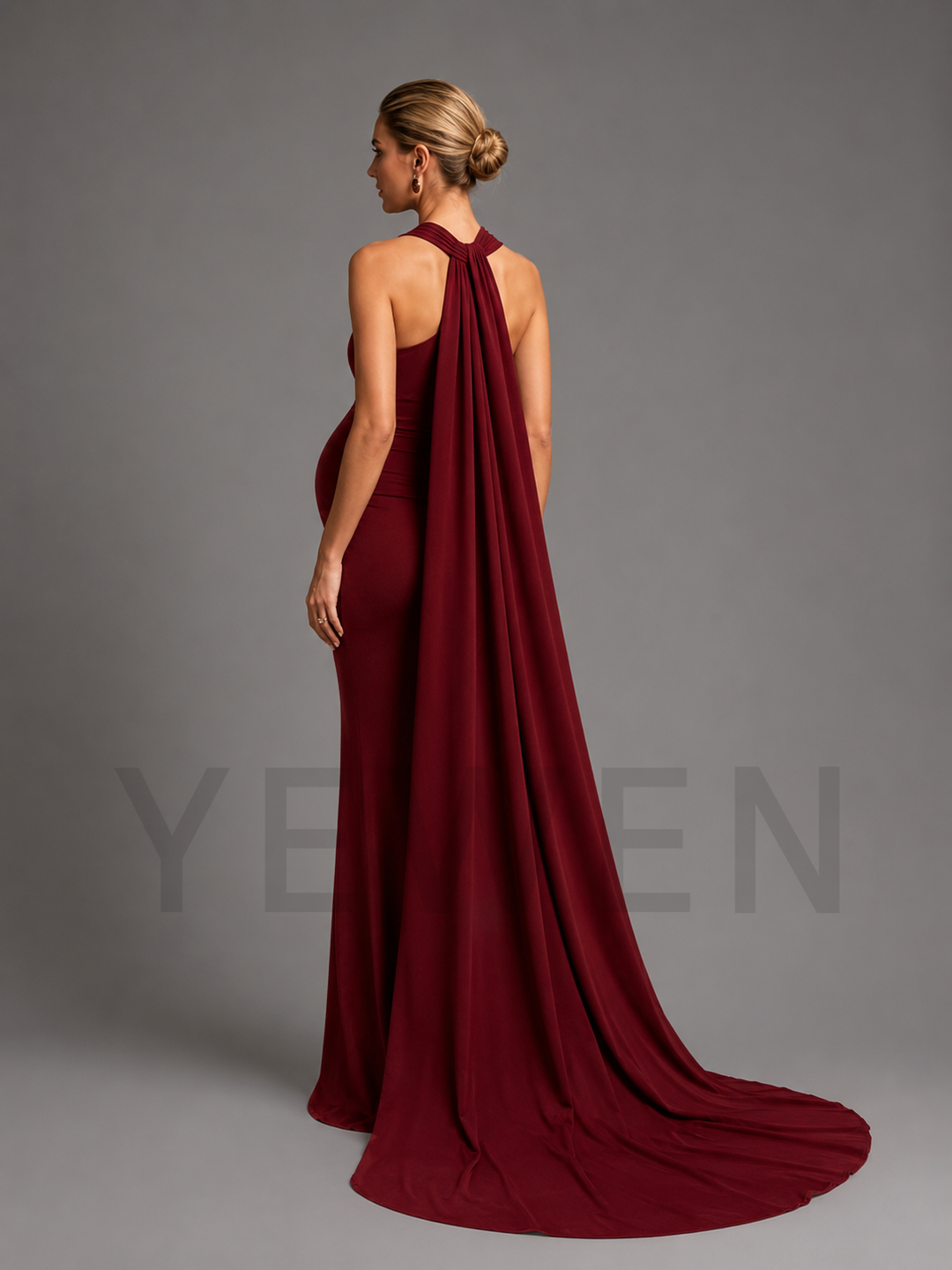 A woman modeling a floor-length burgundy evening gown from the back, featuring a high halter-style neckline with a dramatic draped train, photographed against a neutral gray studio background.