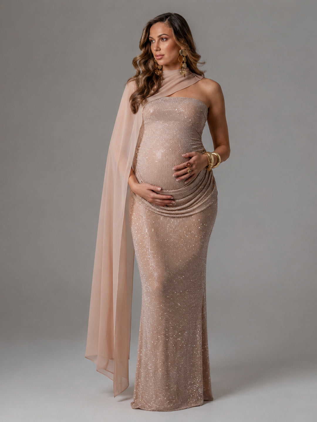 A pregnant woman modeling a champagne-colored, one-shoulder sequined gown with a matching sheer draped cape, set against a solid gray studio background.