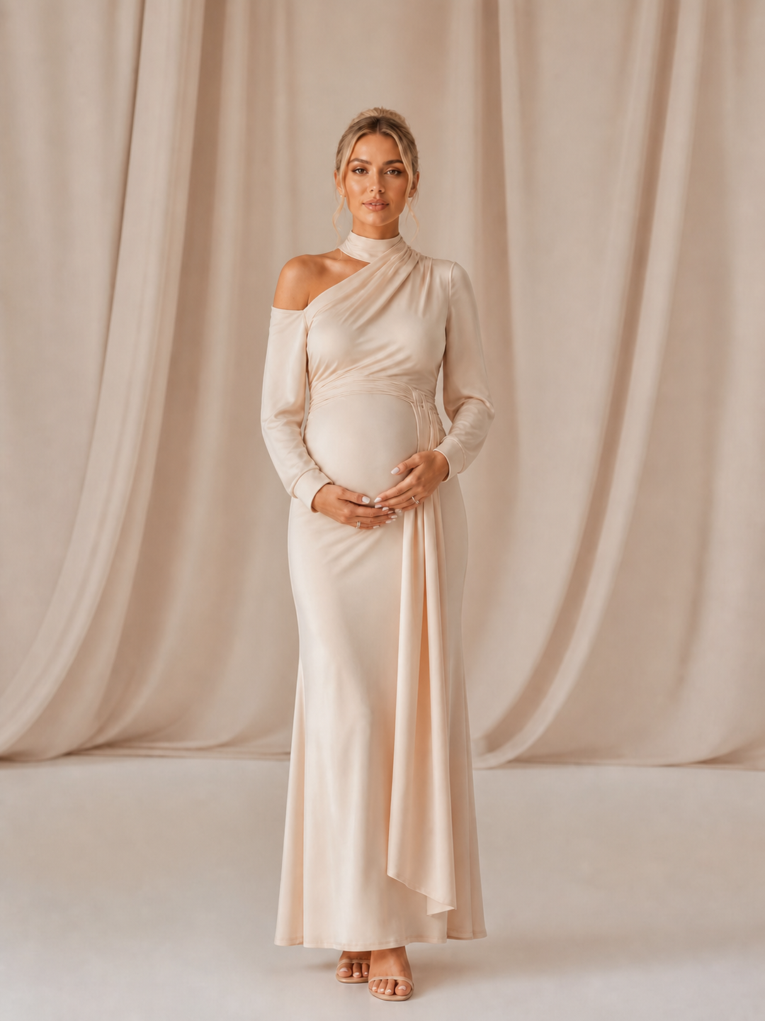 A pregnant woman stands in a studio wearing a long-sleeve, one-shoulder maternity gown in a soft champagne beige color, posed against a backdrop of draped beige fabric.