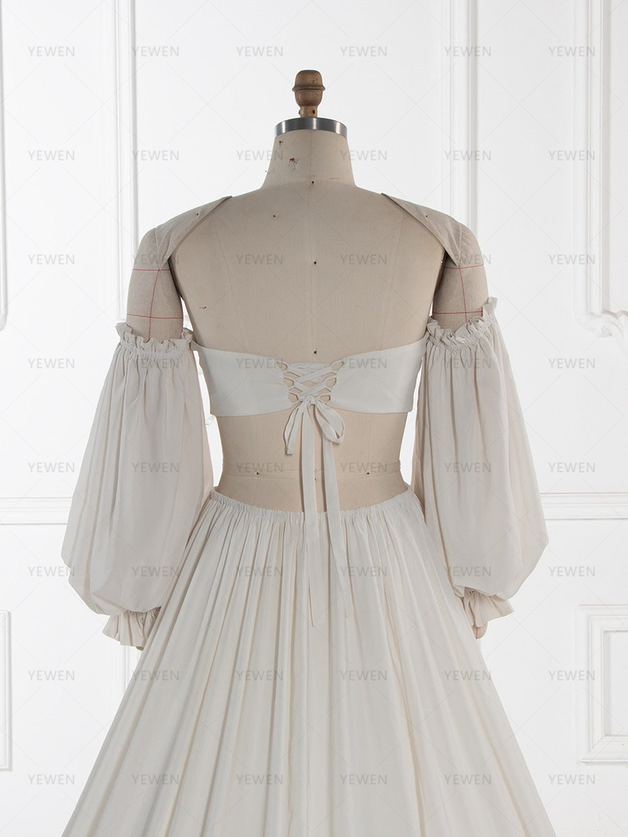 A cream-colored off-the-shoulder dress displayed on a beige dress form mannequin, showing the back lace-up detail and pleated skirt against a white paneled wall background.