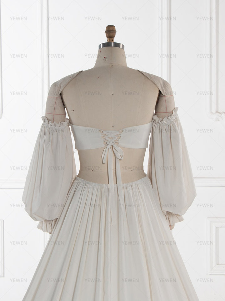 A cream-colored off-the-shoulder dress displayed on a beige dress form mannequin, showing the back lace-up detail and pleated skirt against a white paneled wall background.