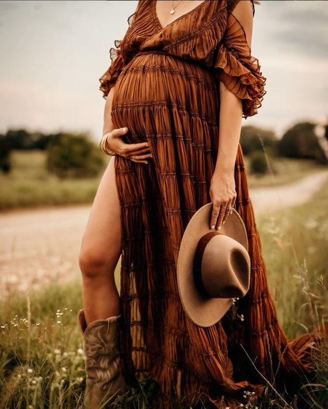 Woman in a long brown dress holding a hat, standing in a field.