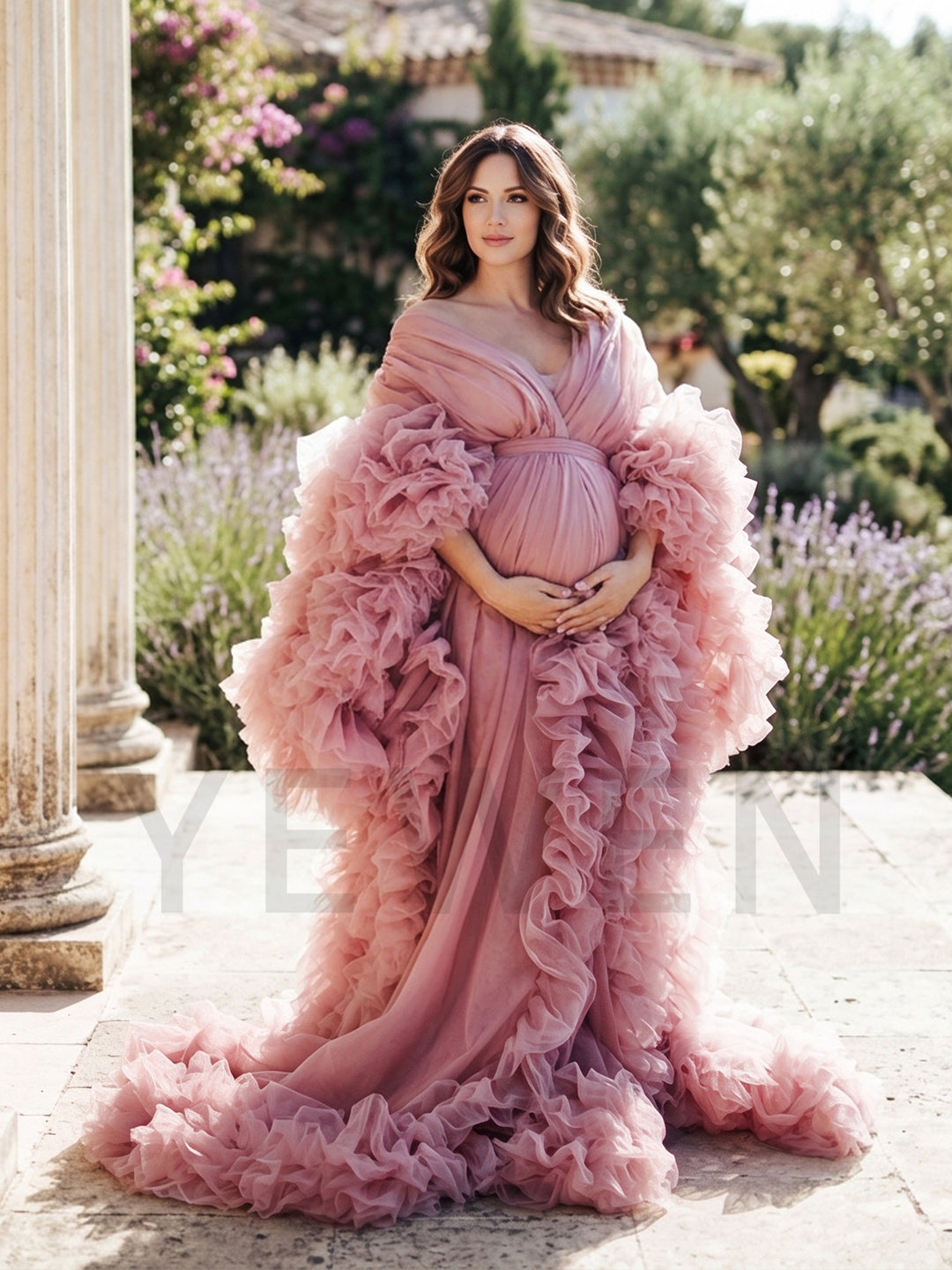 Pregnant woman wearing a dusty pink tulle ruffle sleeve maternity photoshoot gown in an outdoor garden.