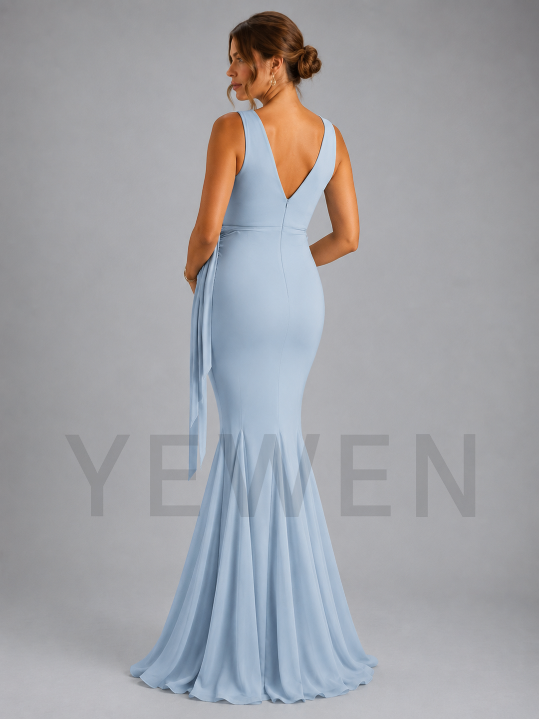 A woman modeling a light blue sleeveless mermaid gown from the back, featuring a deep V-neckline, side tie detail, and a flared skirt, set against a solid gray studio background.