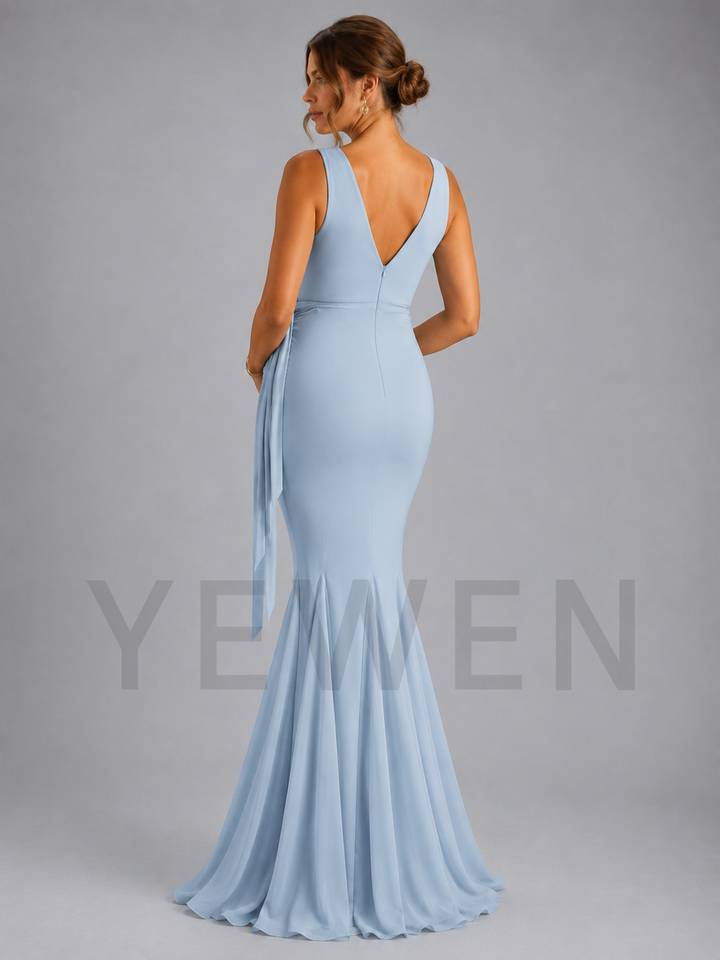 A woman modeling a light blue sleeveless mermaid gown from the back, featuring a deep V-neckline, side tie detail, and a flared skirt, set against a solid gray studio background.
