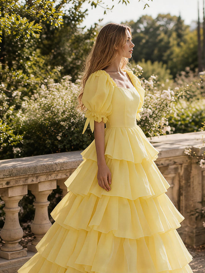 A woman wearing a bright yellow tiered ruffle dress stands outdoors next to a stone balustrade, with green foliage and white flowers in the background.