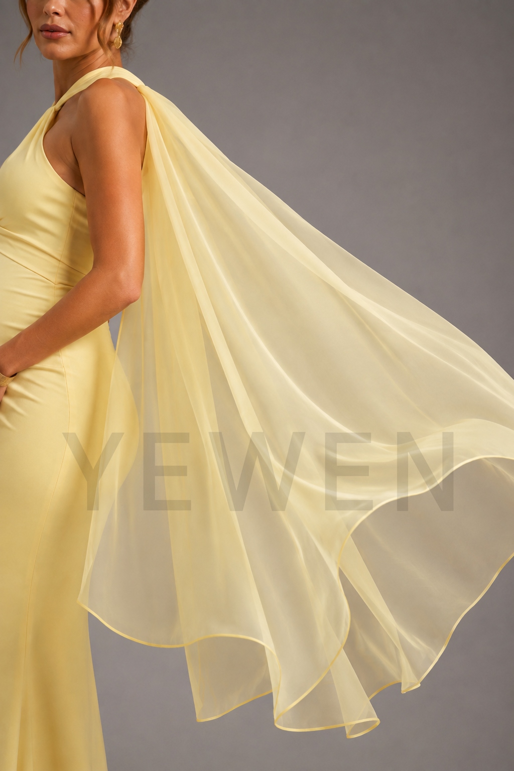 A woman wearing a pale yellow sleeveless gown with a flowing sheer overlay draped across her body, photographed against a plain gray studio background.