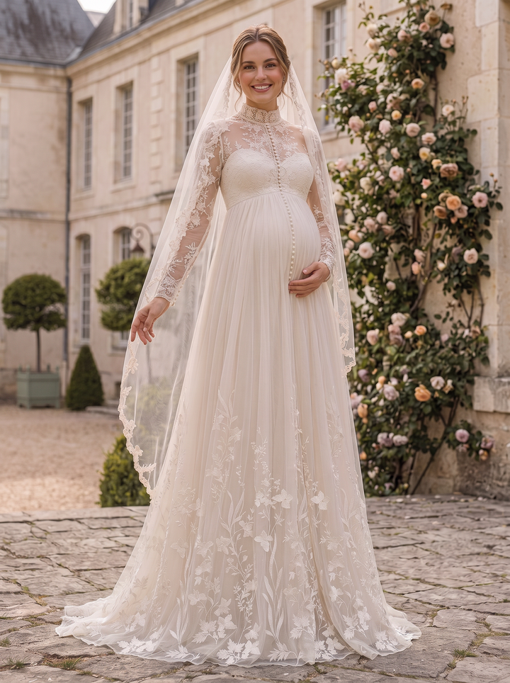 A pregnant woman wearing a long-sleeved ivory lace wedding dress with a matching sheer veil, standing outdoors in front of a stone building with rose vines.