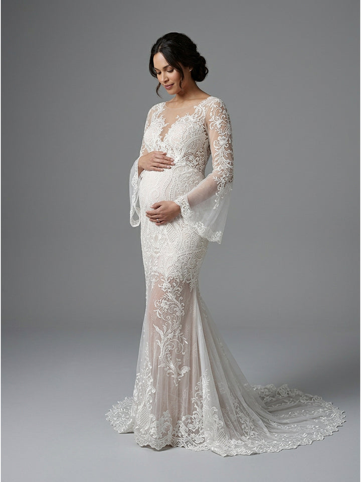 A pregnant model wearing a white lace mermaid-style wedding gown with sheer long bell sleeves and a train, photographed against a plain gray studio background.