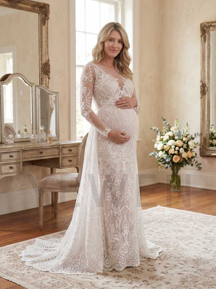 A pregnant woman wearing a long-sleeve lace wedding gown with a deep V-neckline and fitted silhouette, standing in a softly lit bridal room with mirrors, a vanity, and floral arrangements.