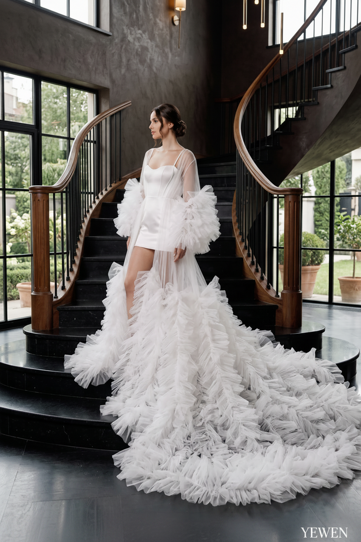 A woman wearing a dramatic white bridal gown with a fitted bodice, sheer draped sleeves, and a voluminous ruffled train, standing on a grand staircase in a modern interior with large windows and dark wood railings.