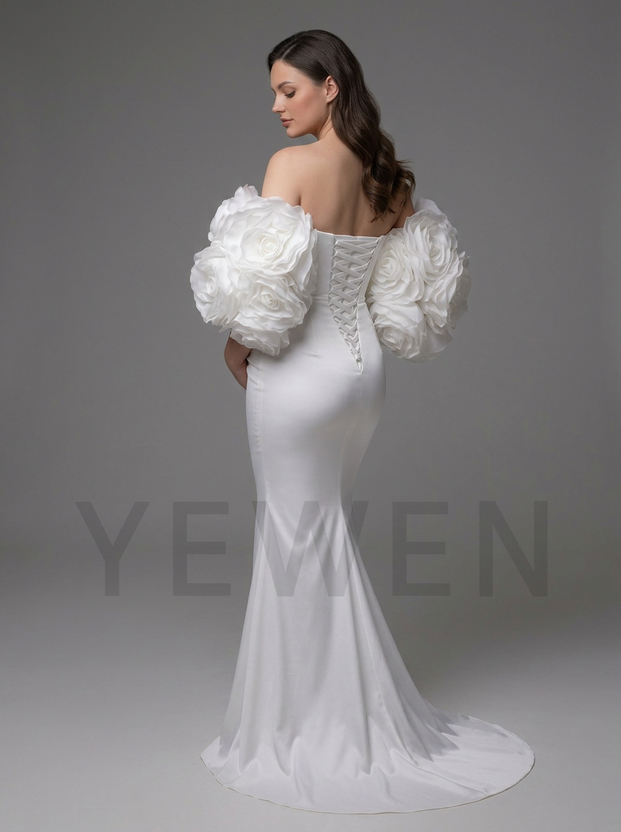 A woman modeling an off-the-shoulder white mermaid gown with dramatic sculptural floral puff sleeves and a lace-up corset-style back, photographed against a smooth gray studio background.