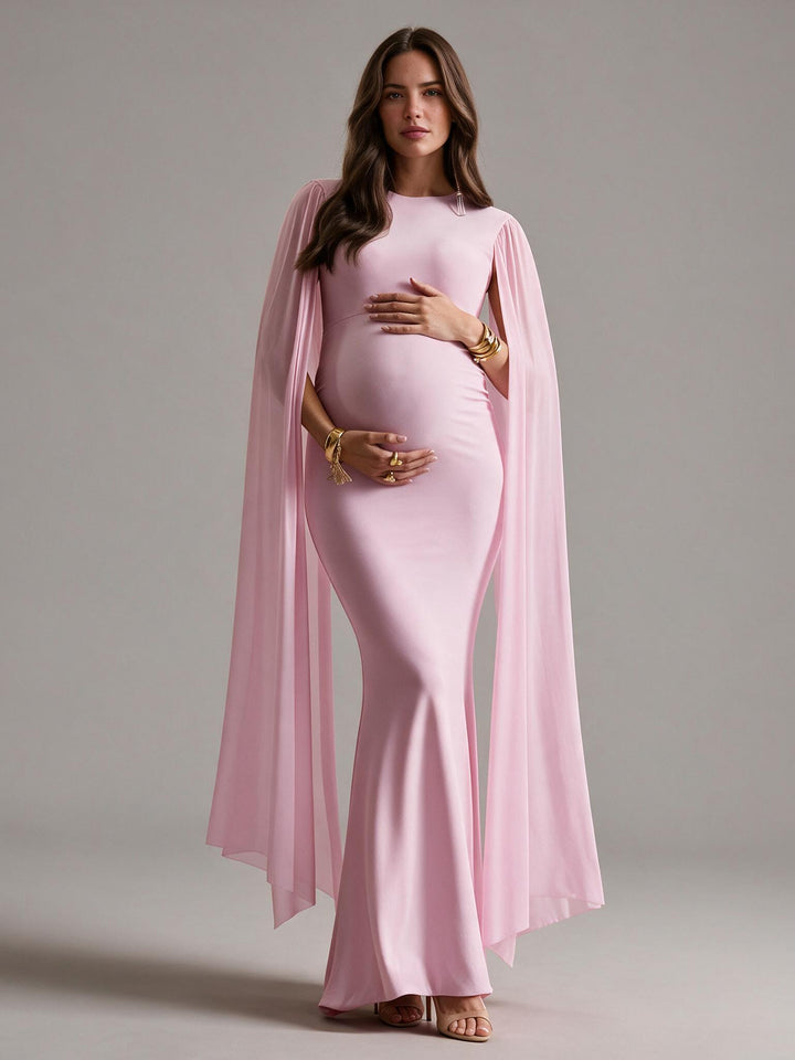 A pregnant woman modeling a light pink maternity gown with a high-neck bodice, fitted silhouette, and flowing sheer cape sleeves, posed against a neutral gray studio background.