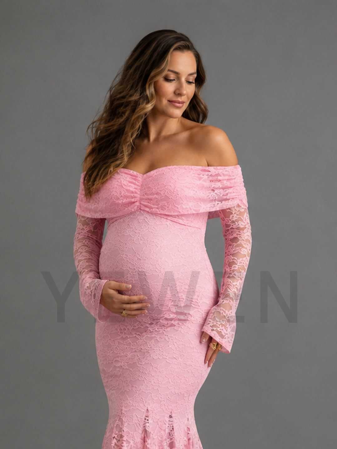 A pregnant model wearing a light pink off-the-shoulder lace maternity gown with long bell sleeves, standing against a solid gray studio background.