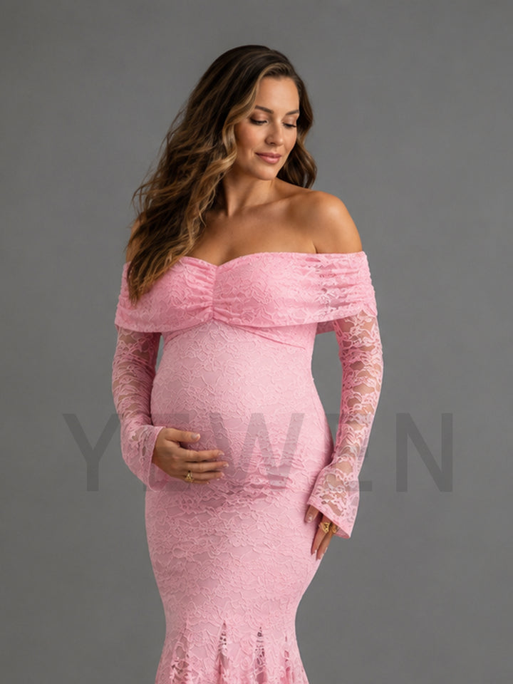 A pregnant model wearing a light pink off-the-shoulder lace maternity gown with long bell sleeves, standing against a solid gray studio background.