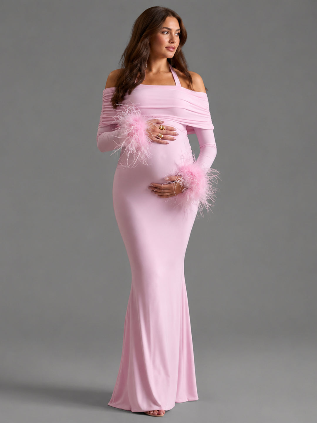 A pregnant model wearing a light pink off-the-shoulder maternity gown with long sleeves and fluffy feather accents on the cuffs, posed against a solid gray studio background.