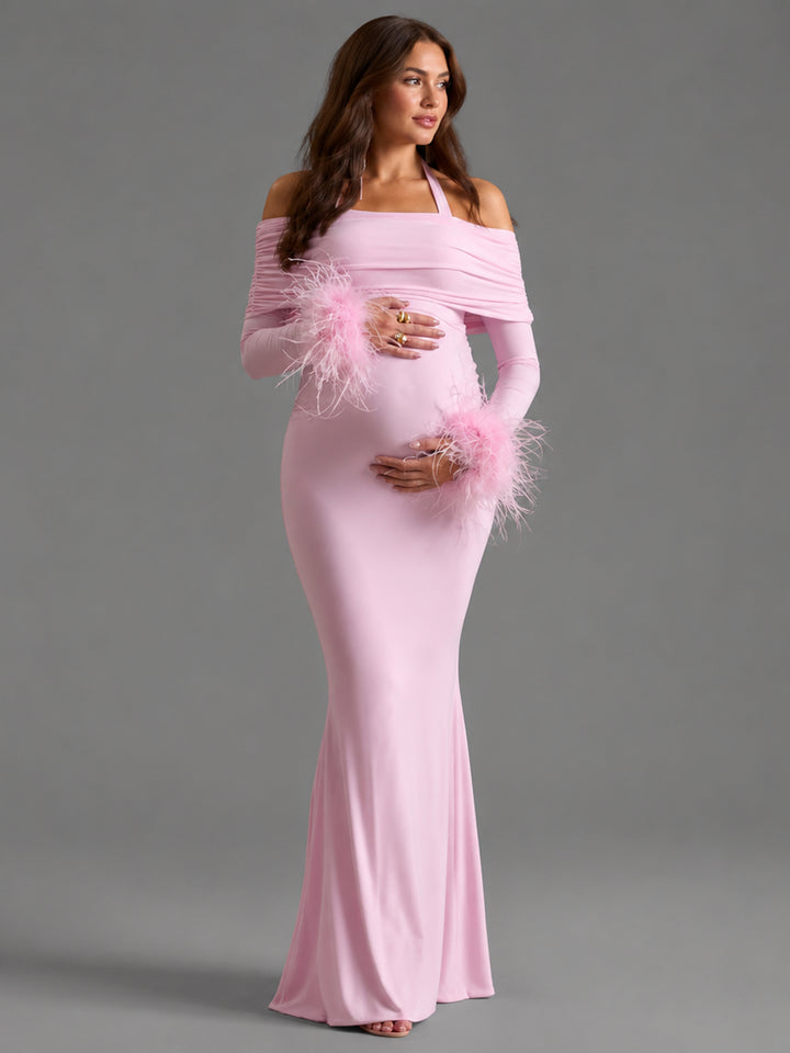 A pregnant model wearing a light pink off-the-shoulder maternity gown with long sleeves and fluffy feather accents on the cuffs, posed against a solid gray studio background.