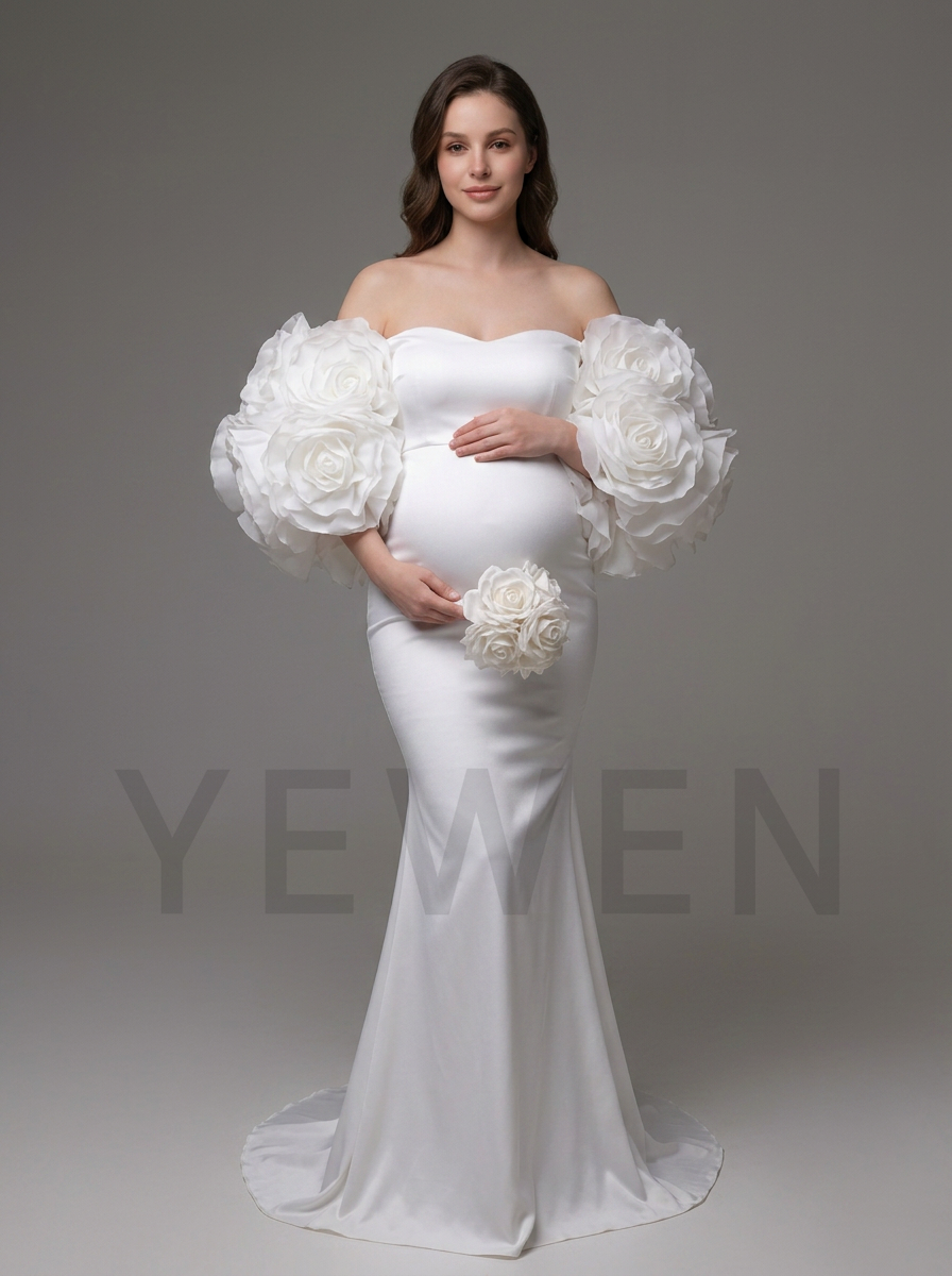 A pregnant model wearing a white off-the-shoulder maternity gown with oversized 3D floral balloon sleeves and holding a matching floral bouquet, set against a solid gray studio background.
